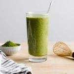 Glass of bright green matcha tea smoothie with bamboo whisk and matcha powder bowl on a wooden table