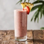 Glass of strawberry and pineapple smoothie topped with fresh fruit, on a rustic table with tropical kitchen background